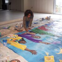 An artist kneels over a mural of women in colorful clothing throwing graduation caps. The artist is painting.