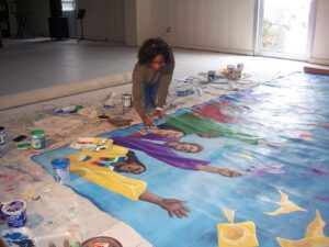 An artist kneels over a mural of women in colorful clothing throwing graduation caps. The artist is painting.