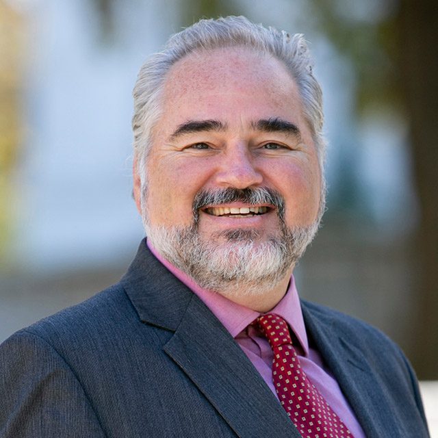 Headshot of a smiling Tom Linfield wearing a dark suit with a red shirt and tie.