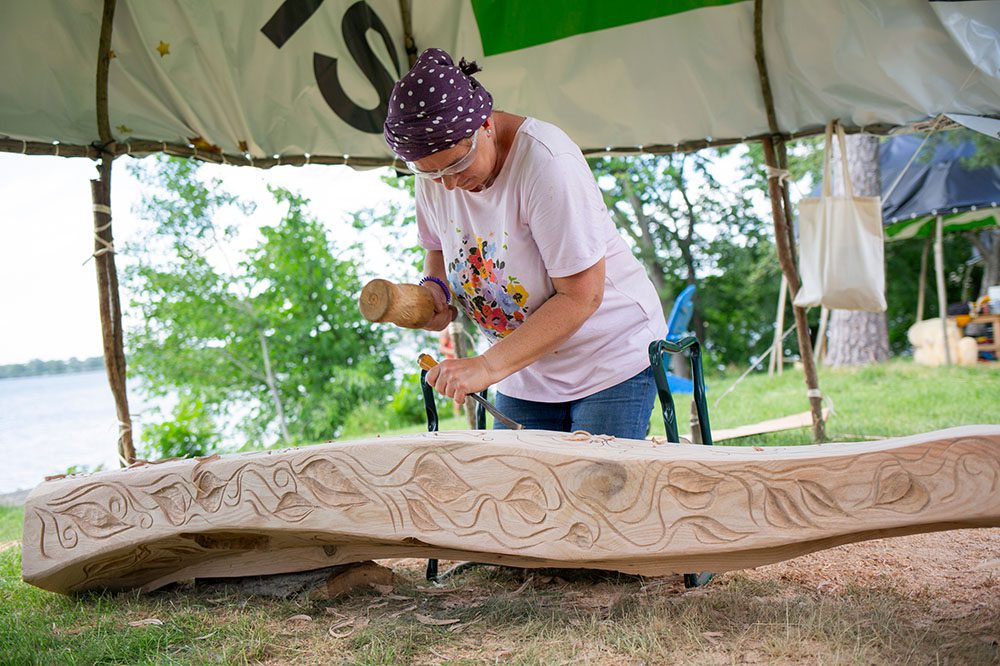 A sculptor holding a chisel works on carving a large wood sculpture slab.