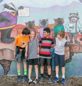 A group of grade school aged boys stands in front of a mural with animal students learning in a classroom.