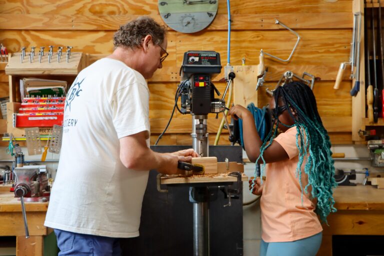 A man assists a young girl with a machine used to drill holes into wood