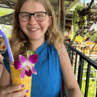 Young girl with glasses smiles as she holds a tall yellow drink with a flower in it