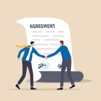 Two men shake hands in front of a signed paper that says 'Agreement'