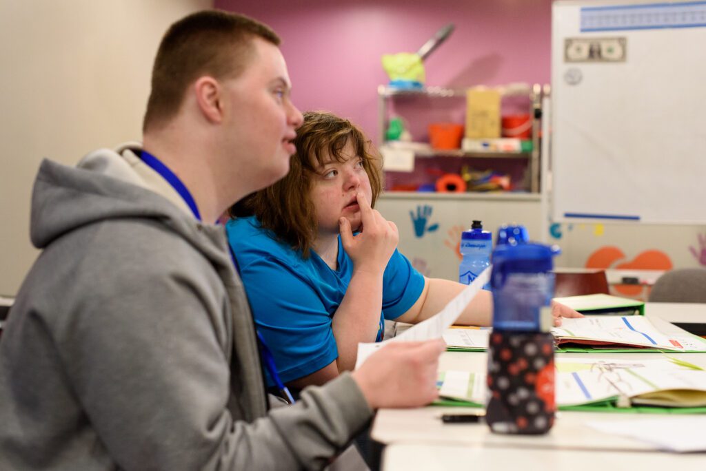 Two individuals with Downs Syndrome look towards their teacher with a worksheet in front of them.